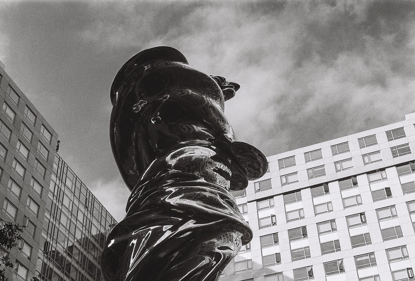 A large, shiny, abstract sculpture stands tall against a cloudy sky, with modern buildings visible on either side.