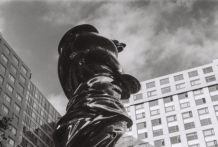 A large, shiny, abstract sculpture stands tall against a cloudy sky, with modern buildings visible on either side.