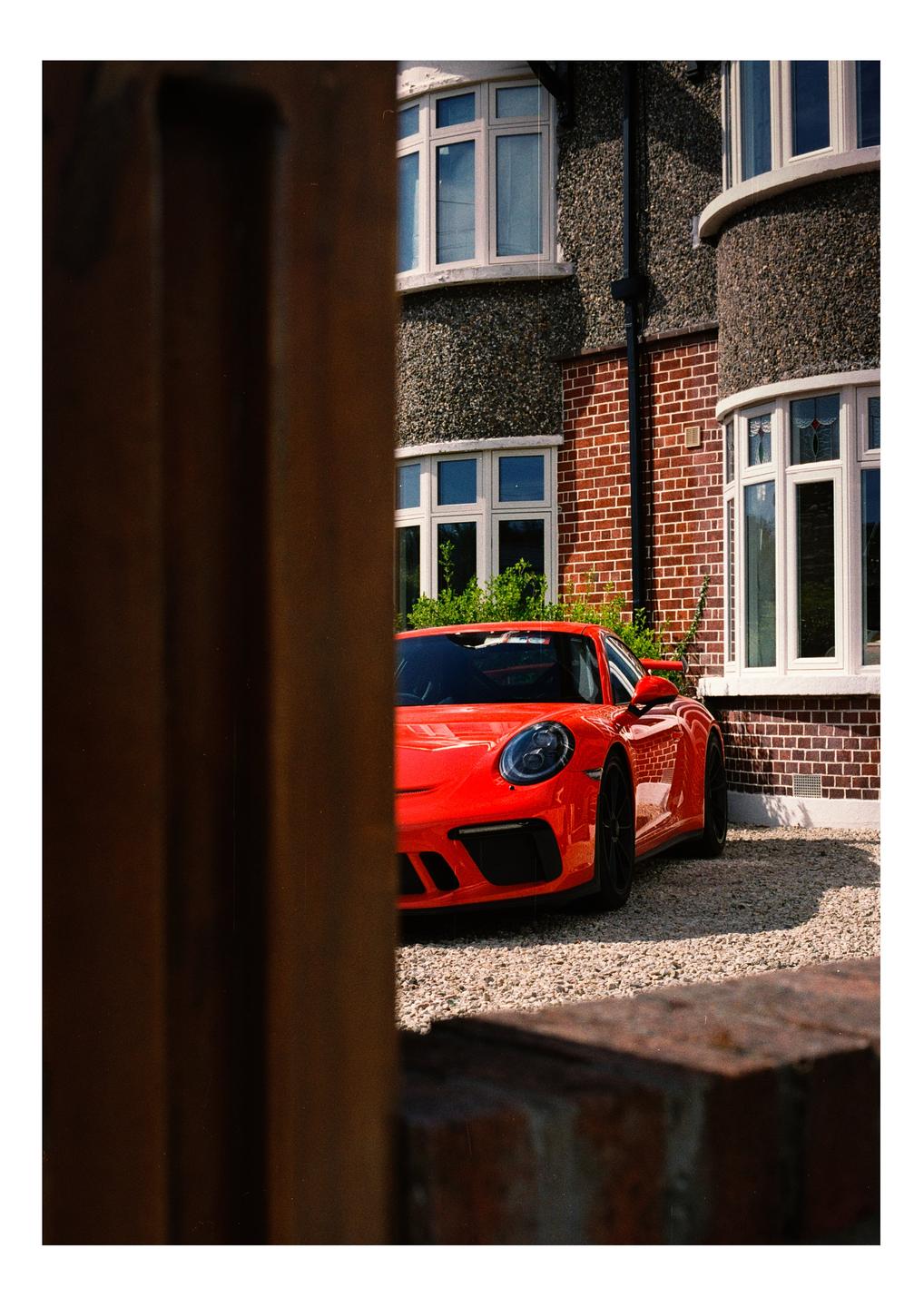 A bright orange sports car is parked on a gravel driveway in front of a brick and stucco house.