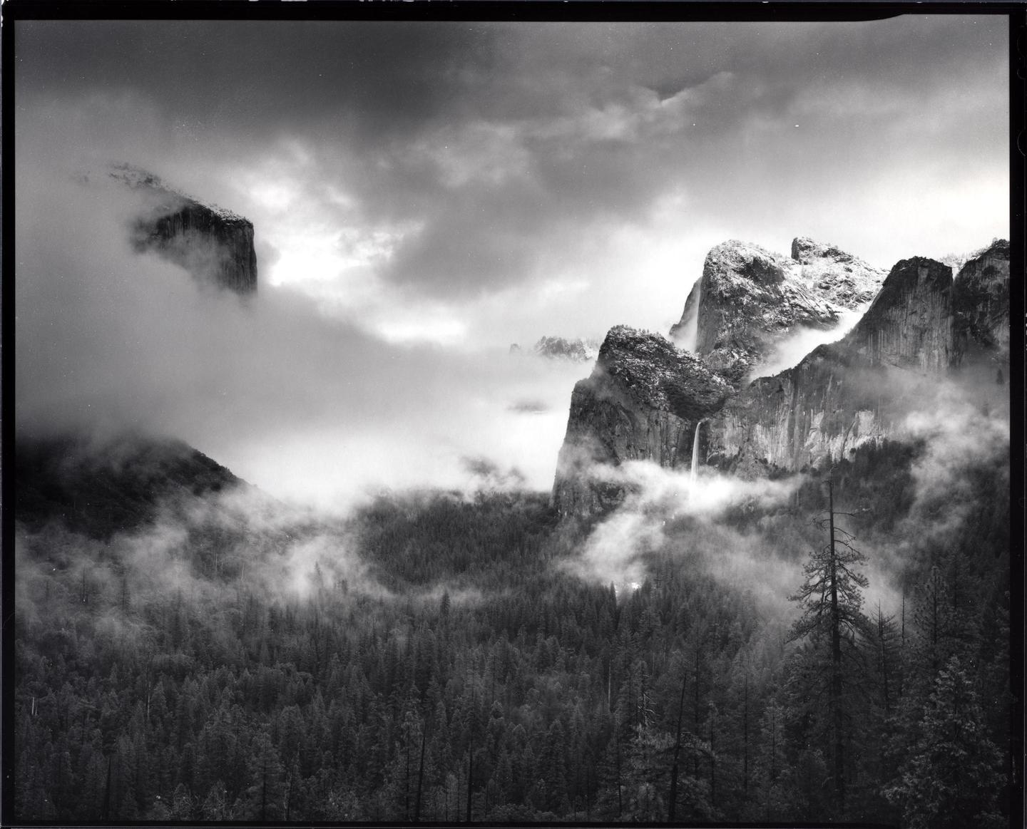 Clouds and fog partially obscure a mountainous landscape with a waterfall and a forest.