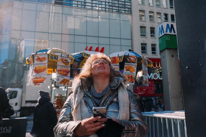 A woman in a silver puffer jacket looks up at the sky while holding a phone in a city street.