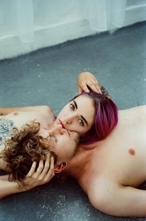 A couple lies on the floor, with the woman resting her head on the man's chest and looking at the viewer.