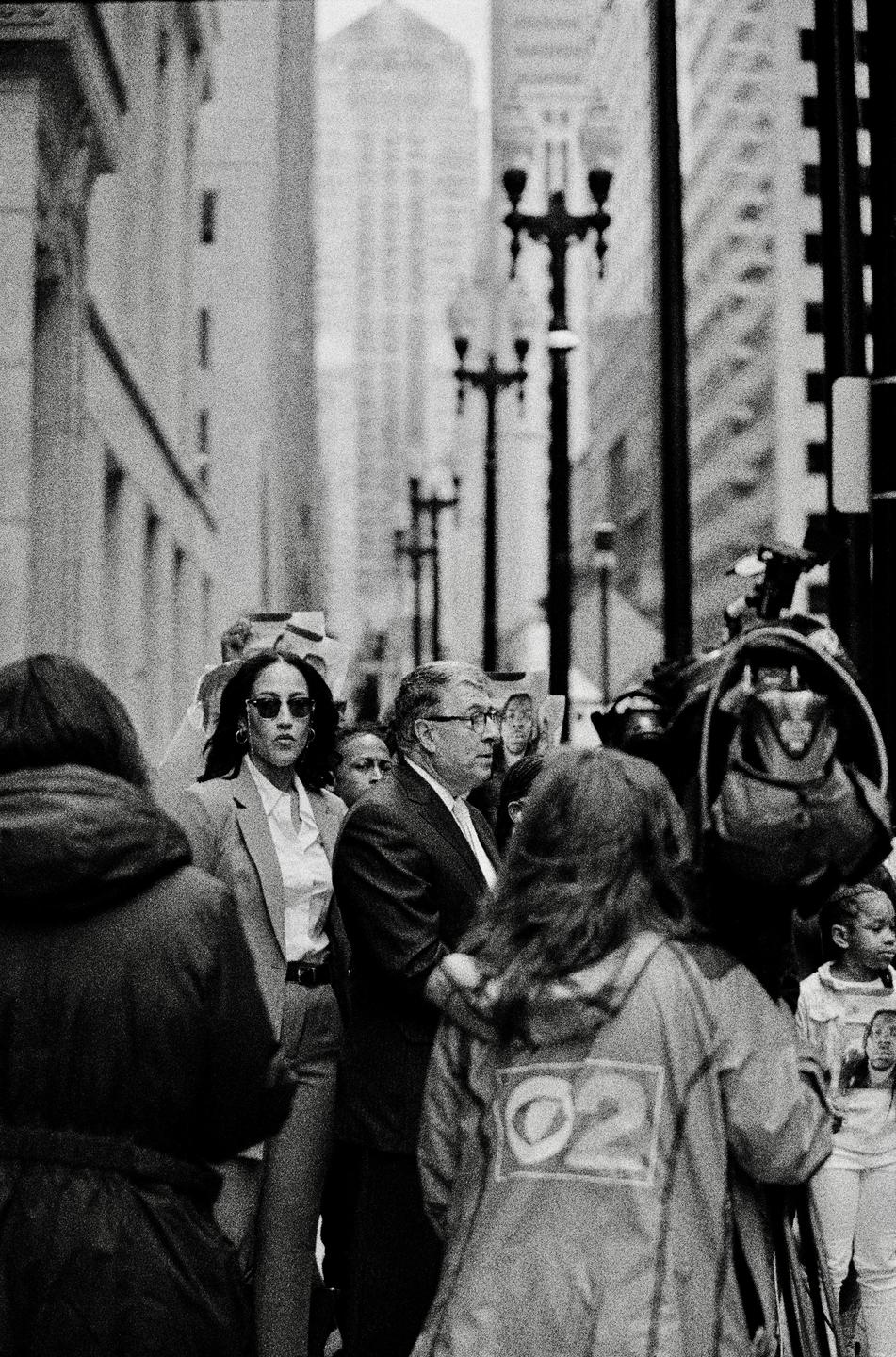 A crowd of people, including a woman in sunglasses and a man in glasses, are gathered on a city street with tall buildings and streetlights in the background.