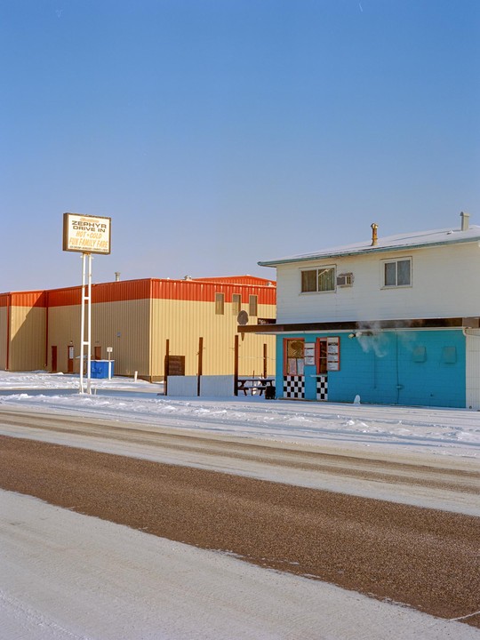 A snow-covered street leads to a drive-in restaurant and a large building under a clear blue sky.