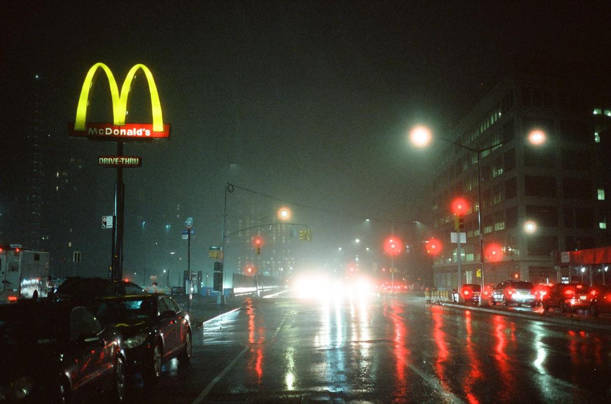 A McDonald's sign stands next to a wet street with cars and buildings at night.