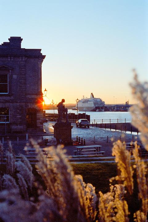 A statue of a man stands on a cobblestone street next to a body of water with a large ship in the distance at sunset.