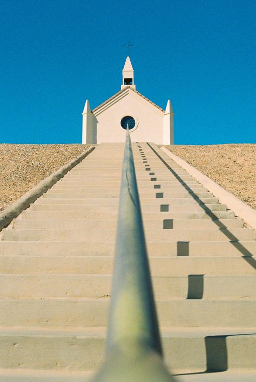A long staircase leads up to a small white church with a round window under a clear blue sky.