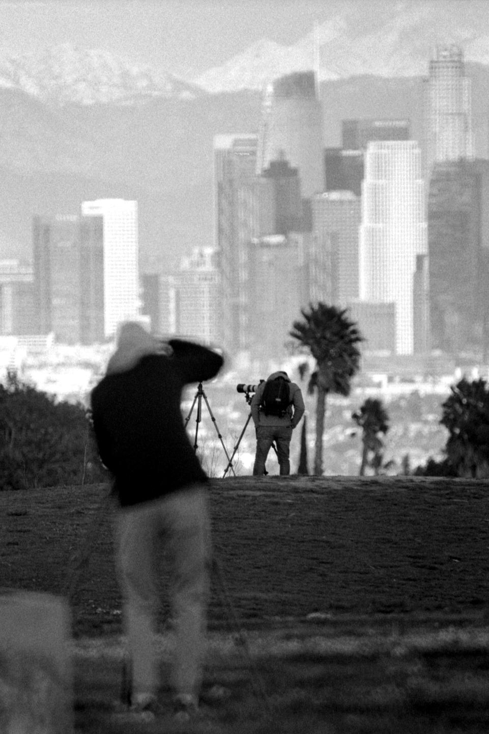 Two men are photographing a distant city skyline with mountains in the background.