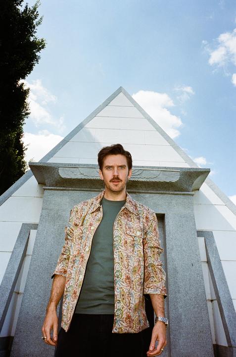 A man with a mustache and a patterned shirt stands in front of a pyramid-shaped structure under a blue sky.