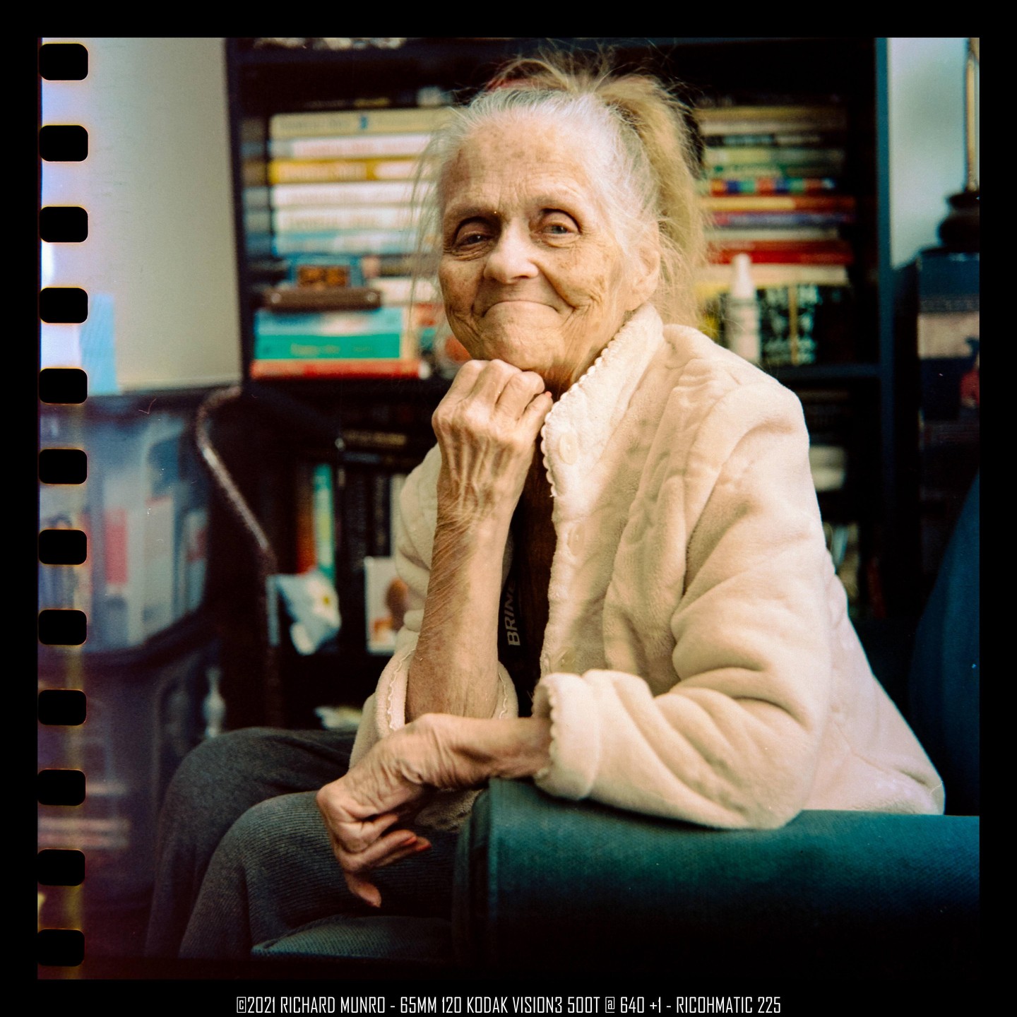 A woman with white hair smiles at the viewer while sitting in a chair with a bookshelf in the background.
