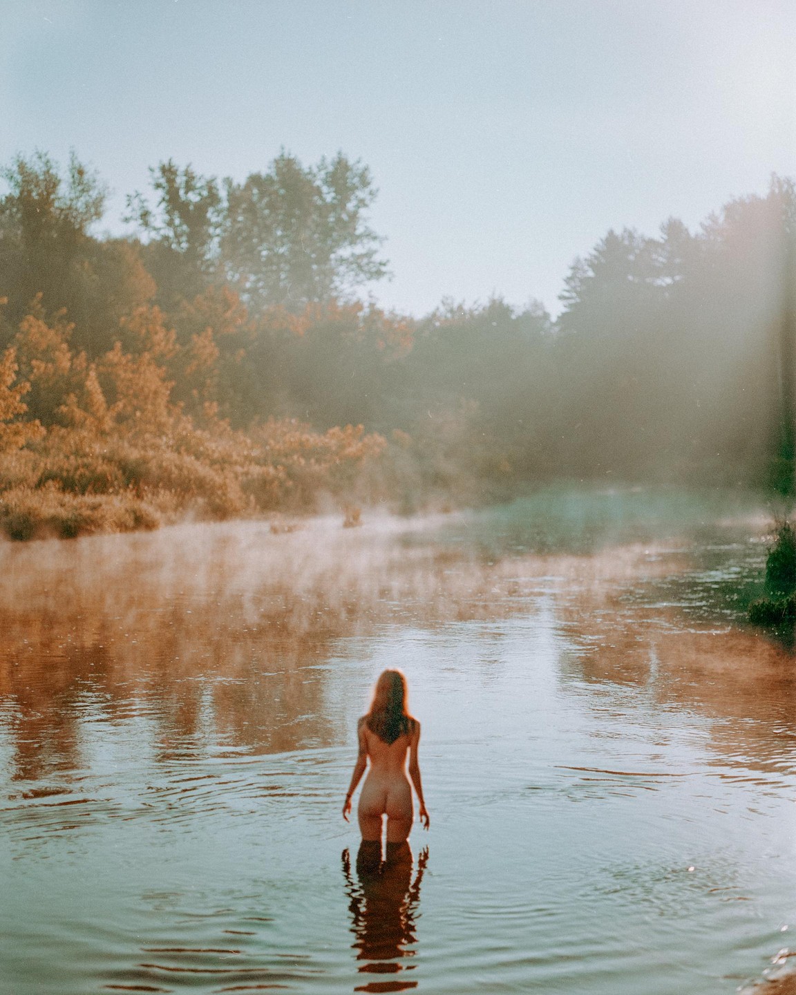 A woman stands in a misty river with trees on the banks.
