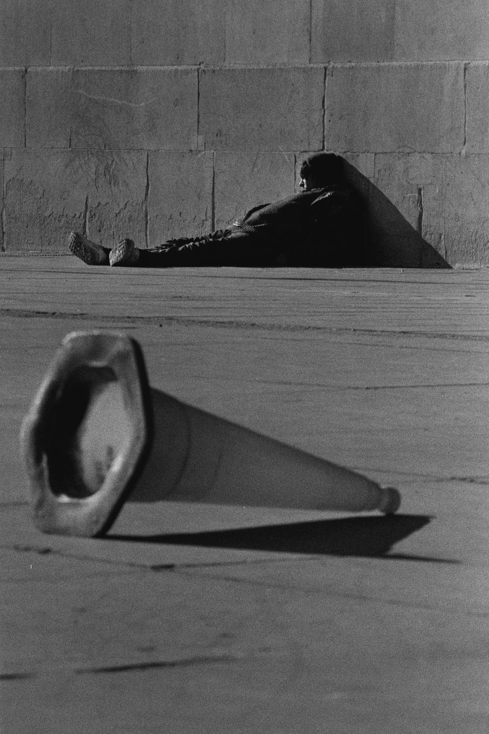 A man rests against a wall while a traffic cone lies on the ground in the foreground.