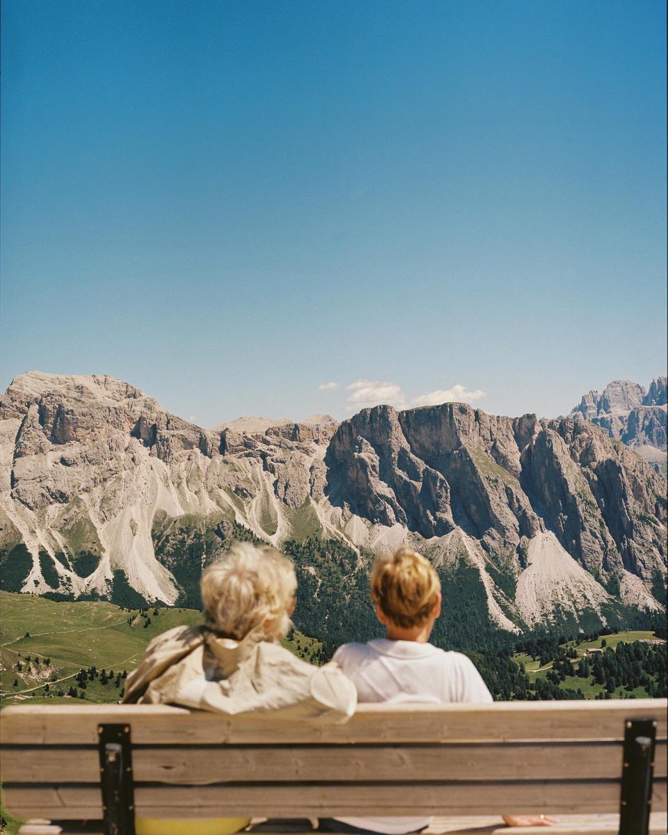 Two women sit on a wooden bench, looking out at a mountain range with green valleys below.