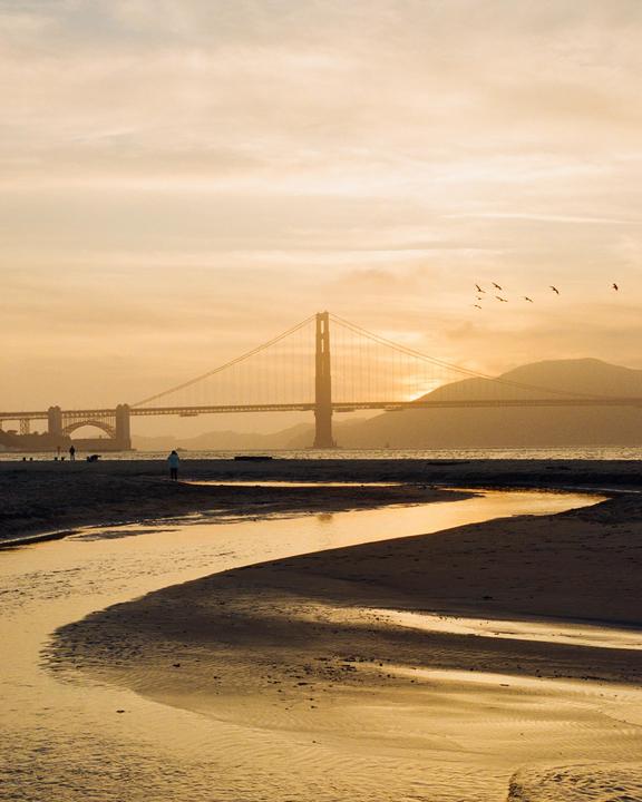 A golden hour view of the Golden Gate Bridge from a sandy beach with a winding stream.