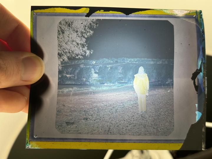 A person in a yellow jacket stands in a field at night, with trees and a body of water in the background.