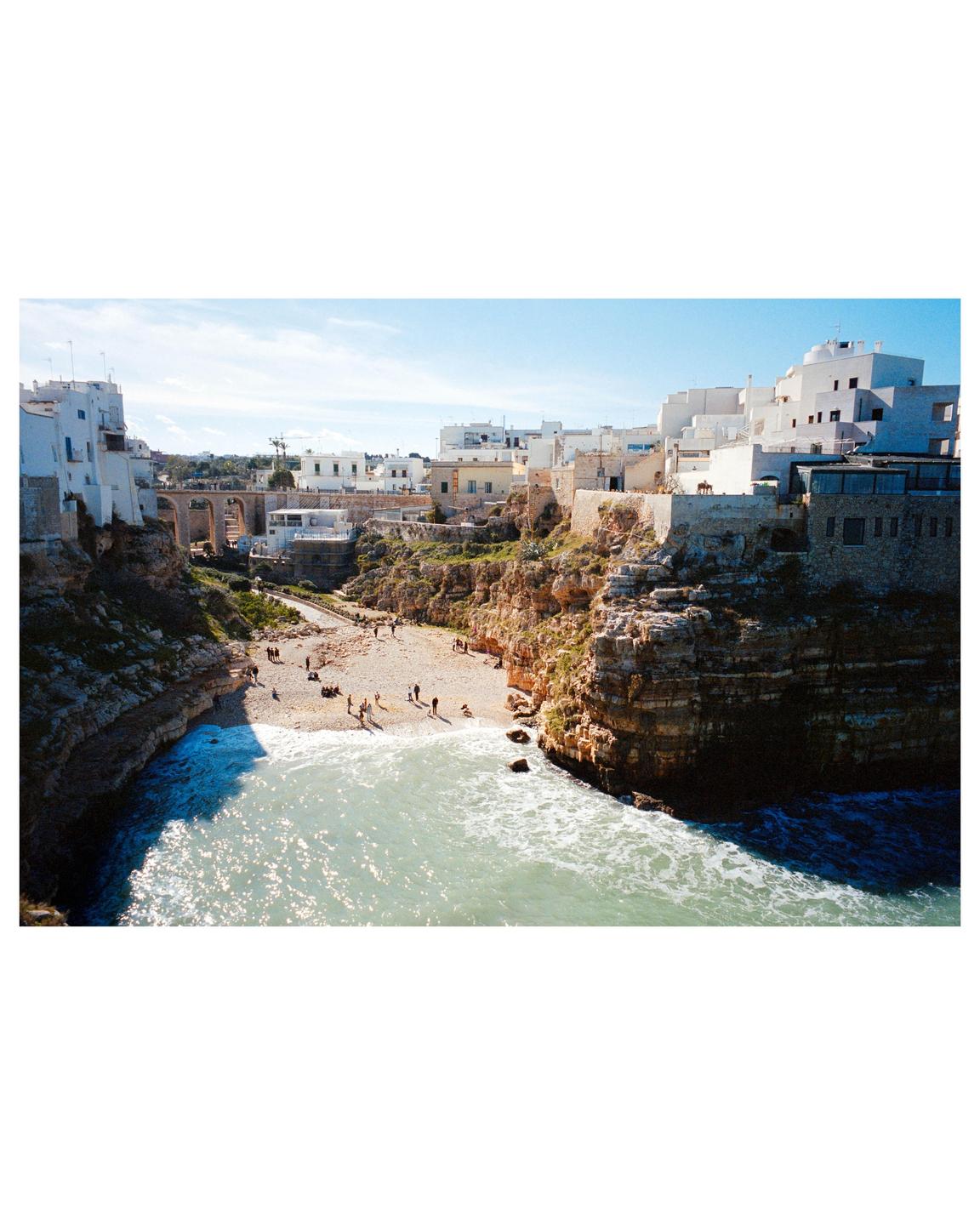 A small beach with people and waves is nestled between cliffs and a town with white buildings.