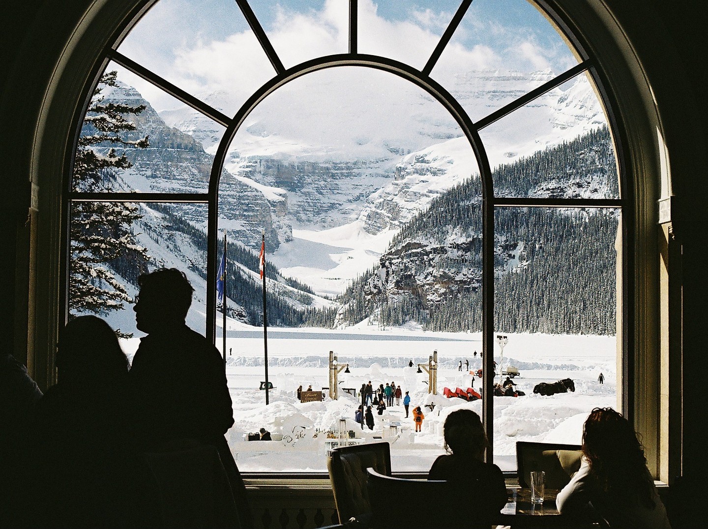 A large arched window frames a snowy mountain landscape with a frozen lake and people enjoying winter activities, while silhouettes of people are visible in the foreground.