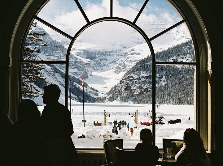 A large arched window frames a snowy mountain landscape with a frozen lake and people enjoying winter activities, while silhouettes of people are visible in the foreground.