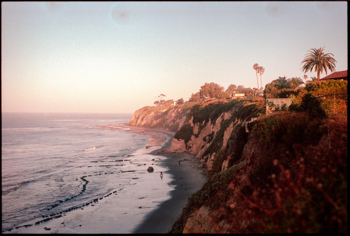 The sun rises over a beach with cliffs and houses on top, with a few people walking on the sand.