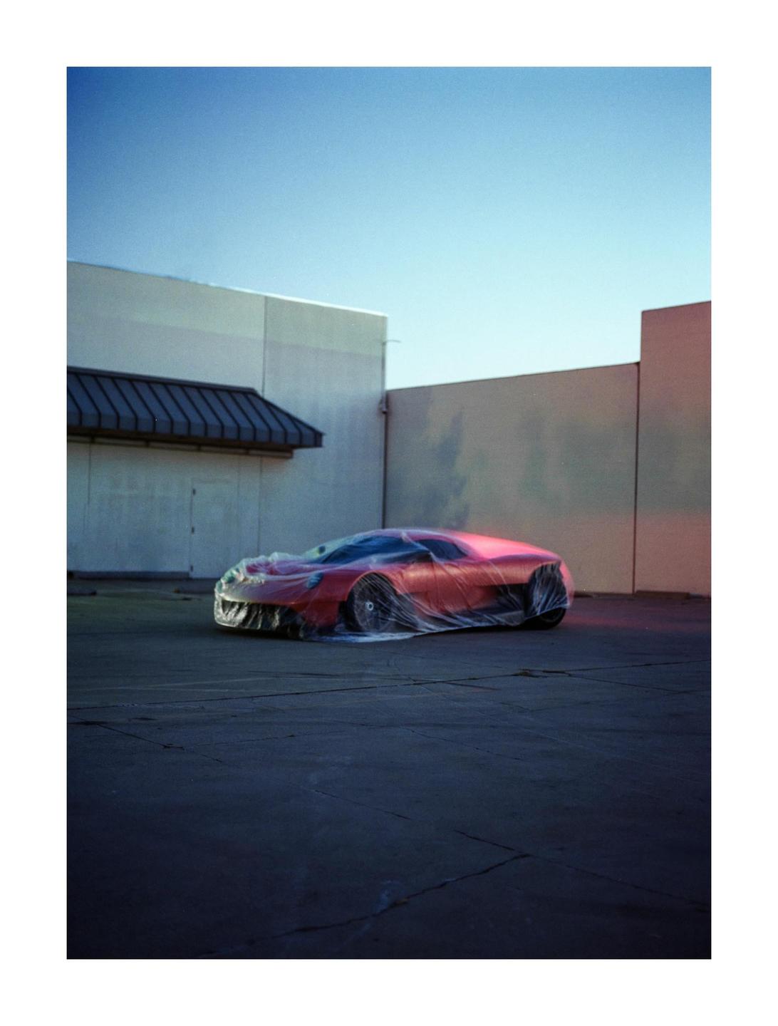 A red concept car covered in plastic is parked in a lot next to a building.