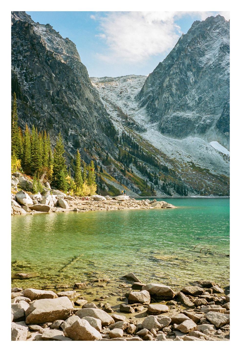 A person stands on the rocky shore of a turquoise lake surrounded by mountains and pine trees.