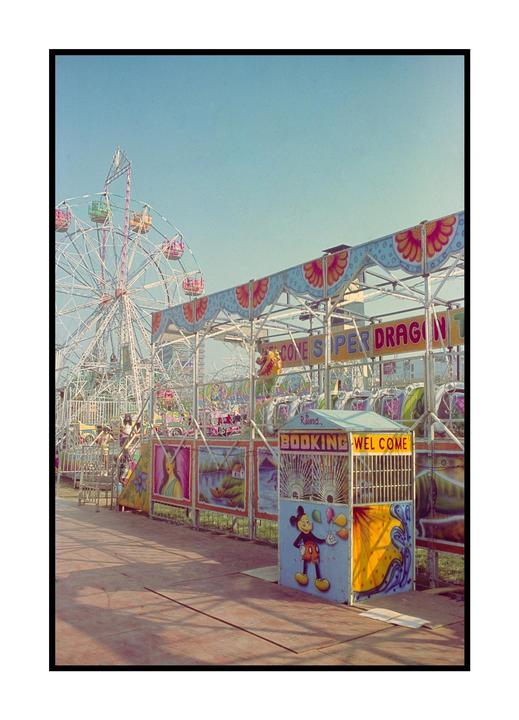 A ferris wheel and a fairground ride are visible under a clear sky, with a booking booth in the foreground.