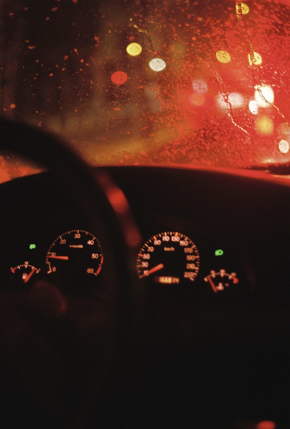 The dashboard of a car is visible through a rain-streaked windshield at night, with blurred red and yellow lights in the background.