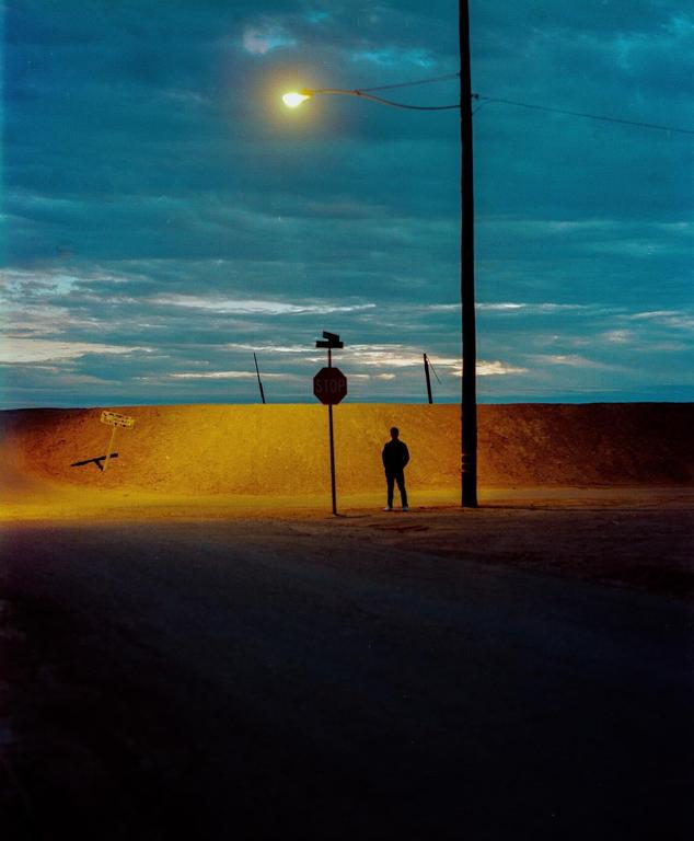 A man stands next to a stop sign and a utility pole under a street light at dusk.