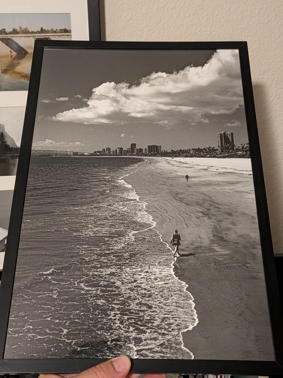 A black and white photograph shows a person walking along a beach with a city skyline in the distance.