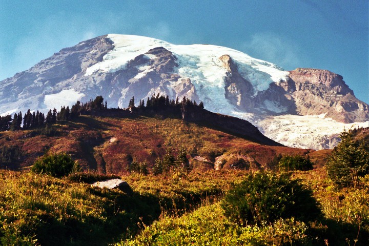 A snow-capped mountain rises above a hillside covered in trees and autumn foliage.