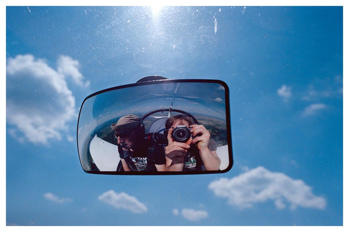 A man takes a self-portrait in a rearview mirror, with another man visible beside him, against a bright blue sky with scattered clouds.