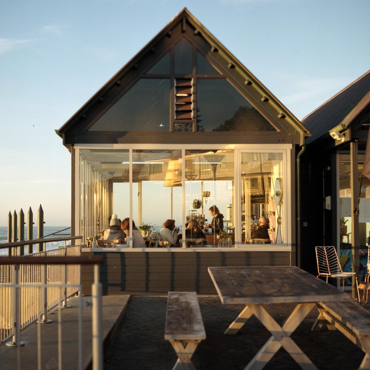 People are sitting inside a cafe with large windows overlooking the ocean at sunset.