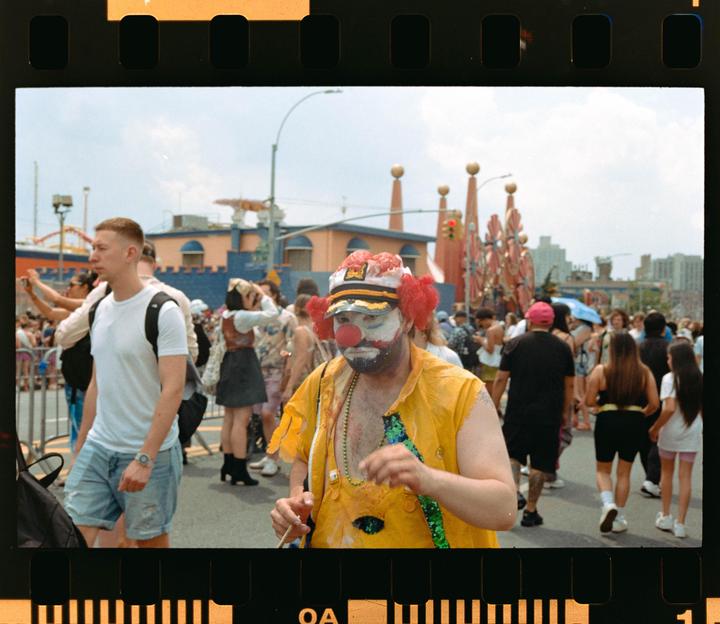 A man dressed as a clown with a red nose and a captain's hat walks through a crowded street during a parade.