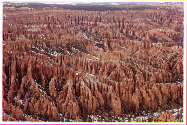 A vast landscape of red rock formations with patches of snow and scattered trees stretches under a distant forest.