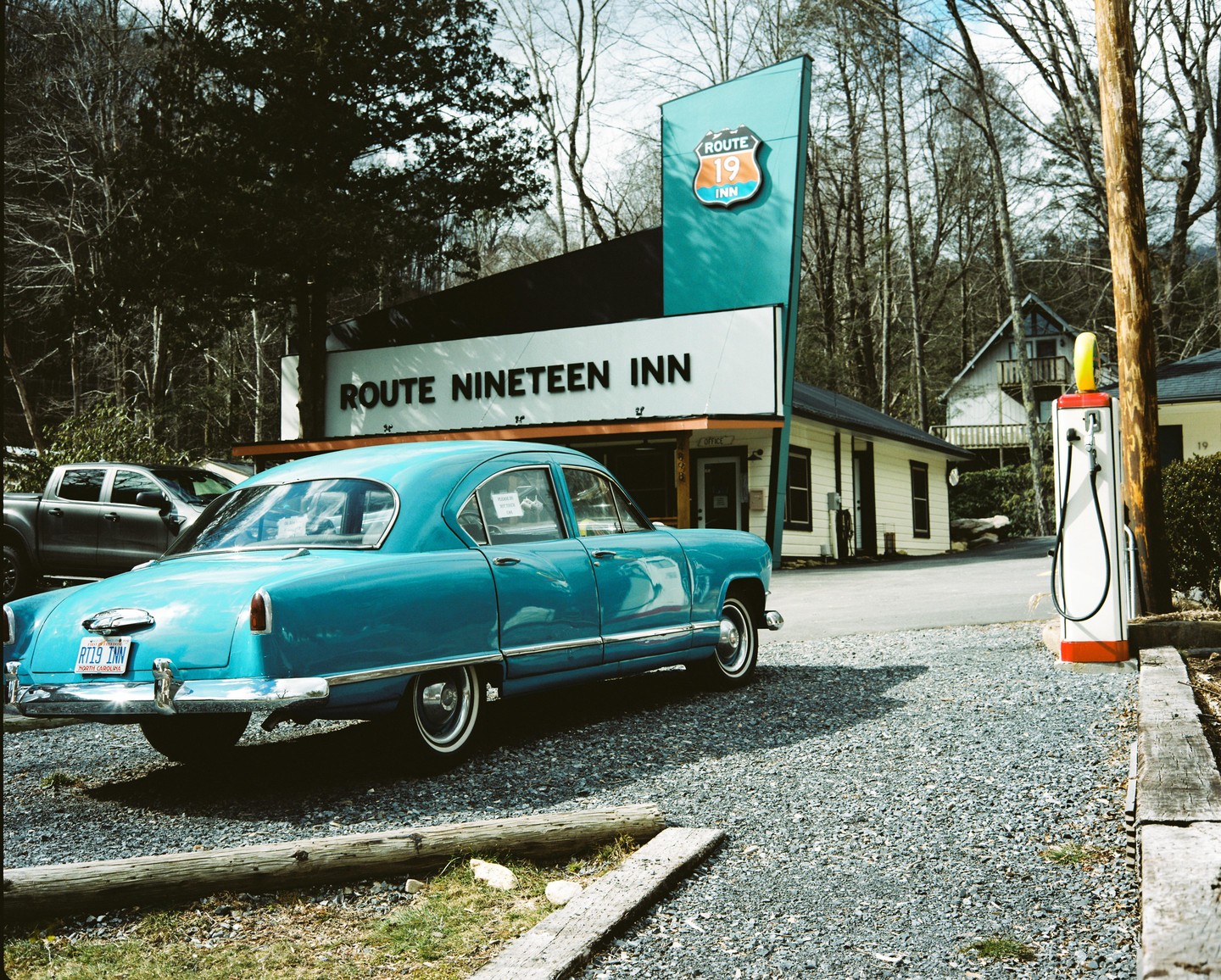 A turquoise vintage car is parked on a gravel lot in front of the Route Nineteen Inn.