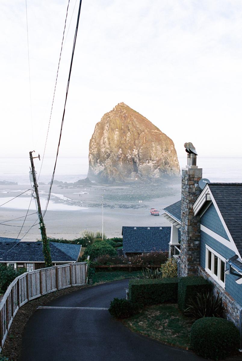 A large rock formation rises from the ocean behind houses and a road.