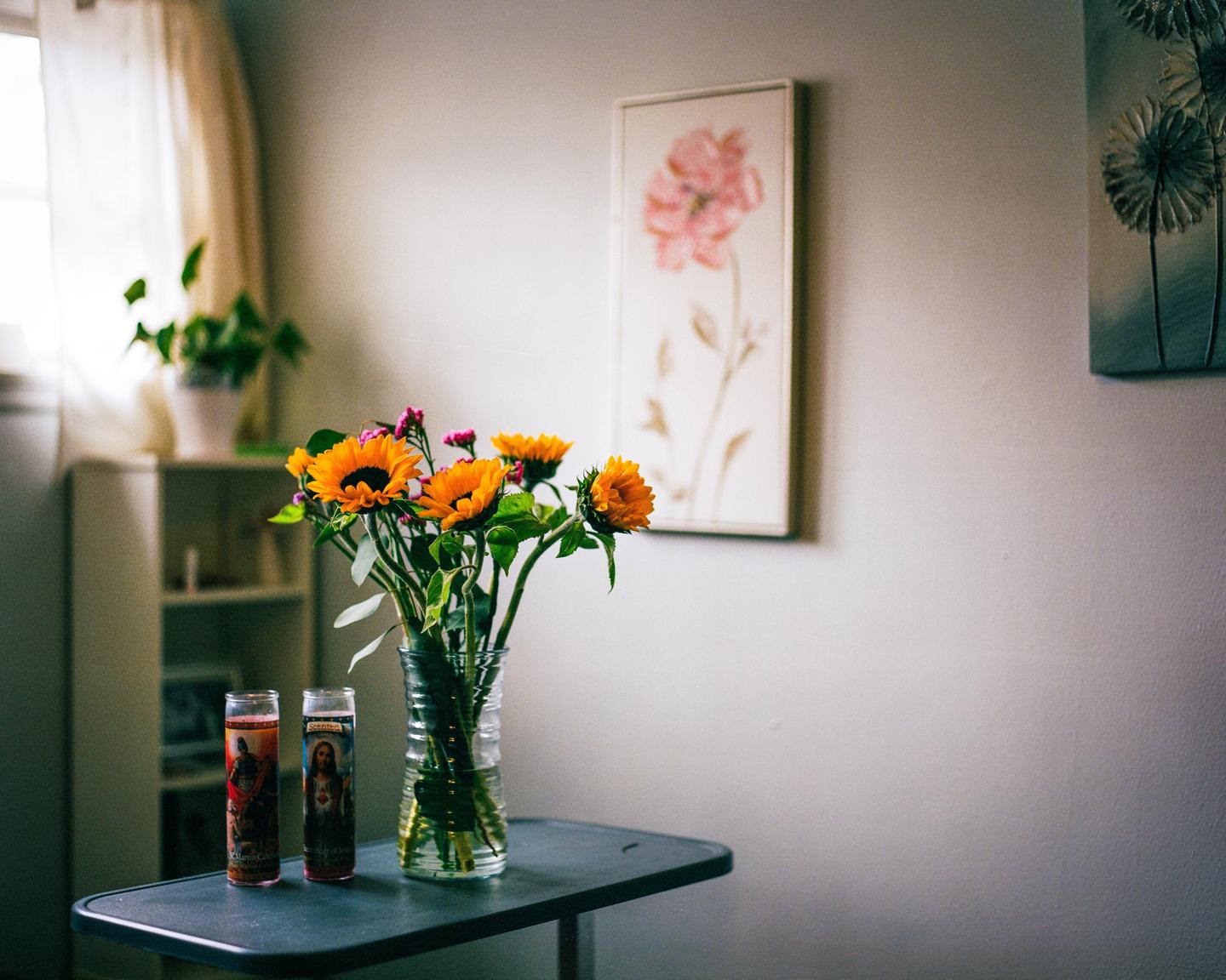 A vase of sunflowers and two religious candles sit on a table in a room with artwork on the wall.