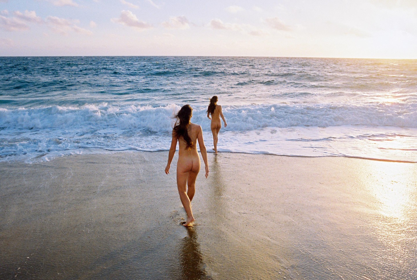 Two women walk into the ocean from a sandy beach at sunset.