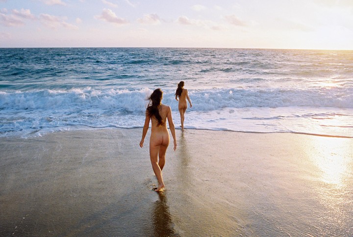 Two women walk into the ocean from a sandy beach at sunset.