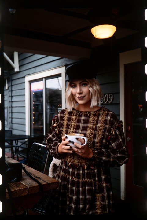 A woman in a plaid dress and hat holds a mug of coffee while standing outside a building with the number 509.