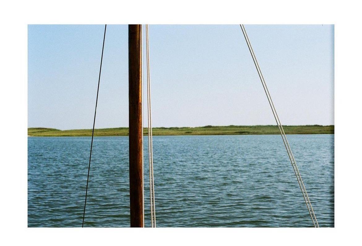 A wooden mast and ropes are visible in the foreground of a body of water with a grassy bank in the distance.