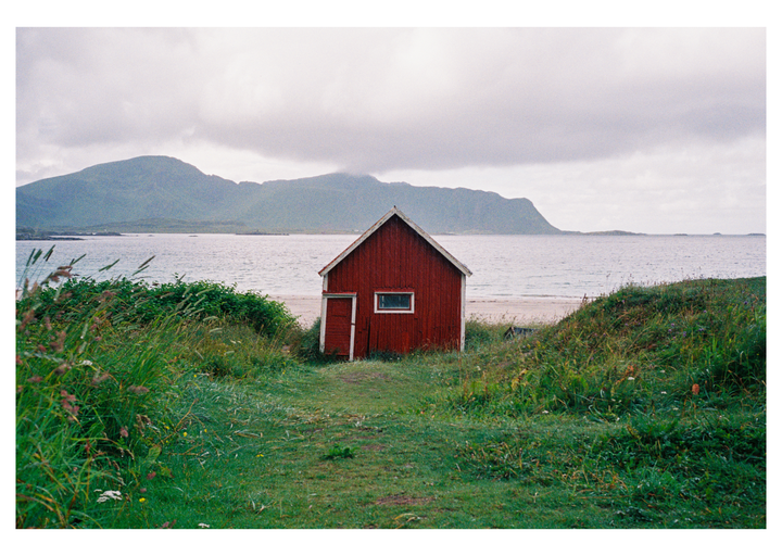 A red cabin sits on a grassy dune overlooking a beach and the sea with mountains in the distance under a cloudy sky.