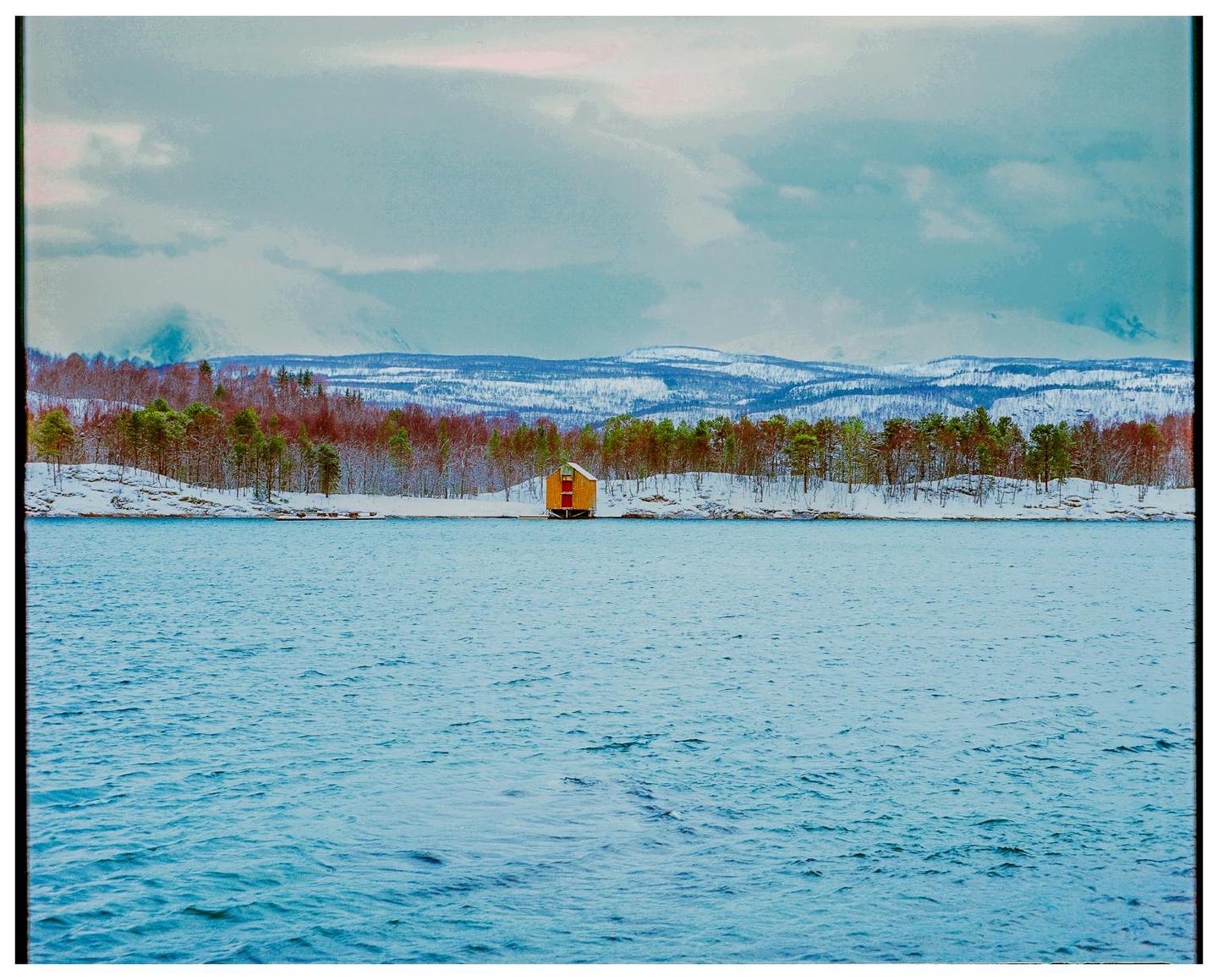 A small yellow house sits on a snowy shoreline with a forest and mountains in the background under a cloudy sky.