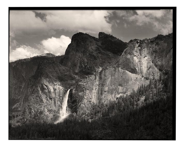 A waterfall cascades down a rocky mountain face with a forest at its base under a cloudy sky.