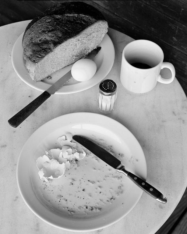 A half loaf of bread, an egg, a salt shaker, and a mug are on a table next to a dirty plate with eggshells and a knife.