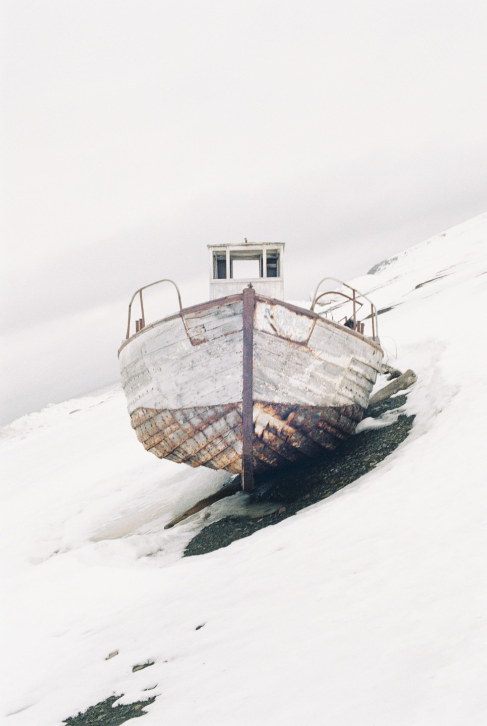 An old, weathered boat rests on a snowy slope with a rocky patch visible beneath its hull.
