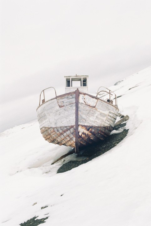 An old, weathered boat rests on a snowy slope with a rocky patch visible beneath its hull.