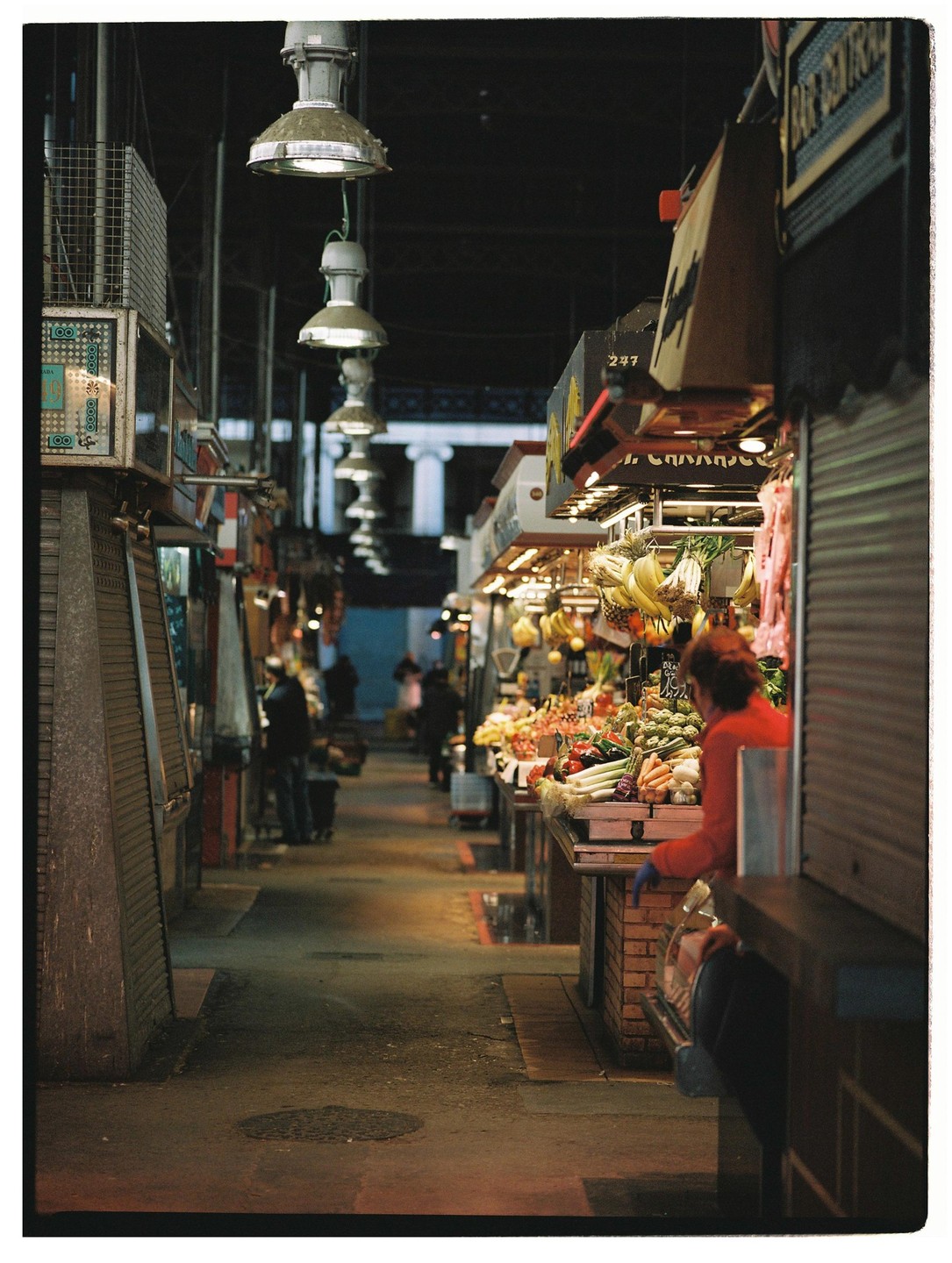 A woman in a red top stands at a market stall filled with produce under hanging lights.