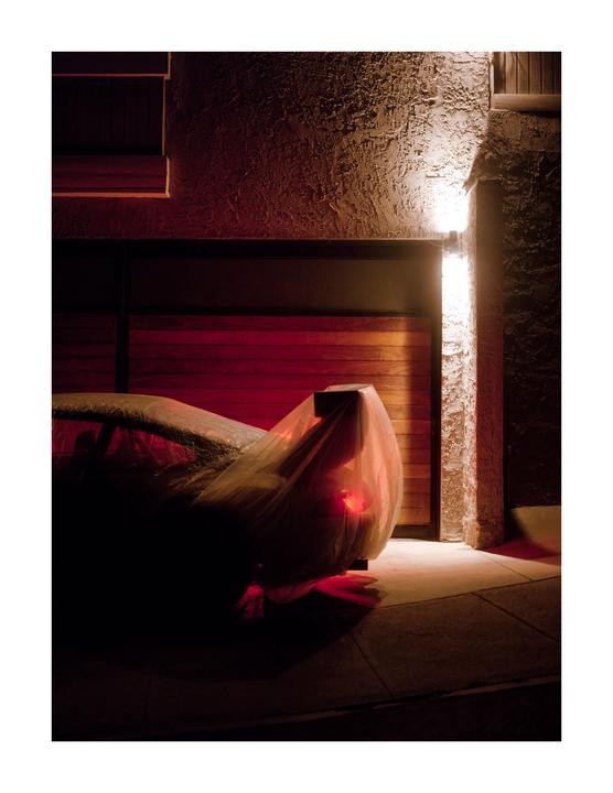 A car covered with a translucent sheet is parked in front of a wooden garage door at night, illuminated by a wall light and a red taillight.