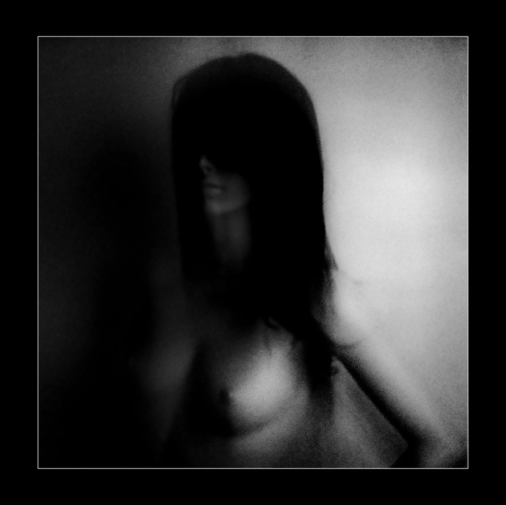 A blurry, high-contrast black and white image shows a woman with long hair covering her face, with her bare torso visible.